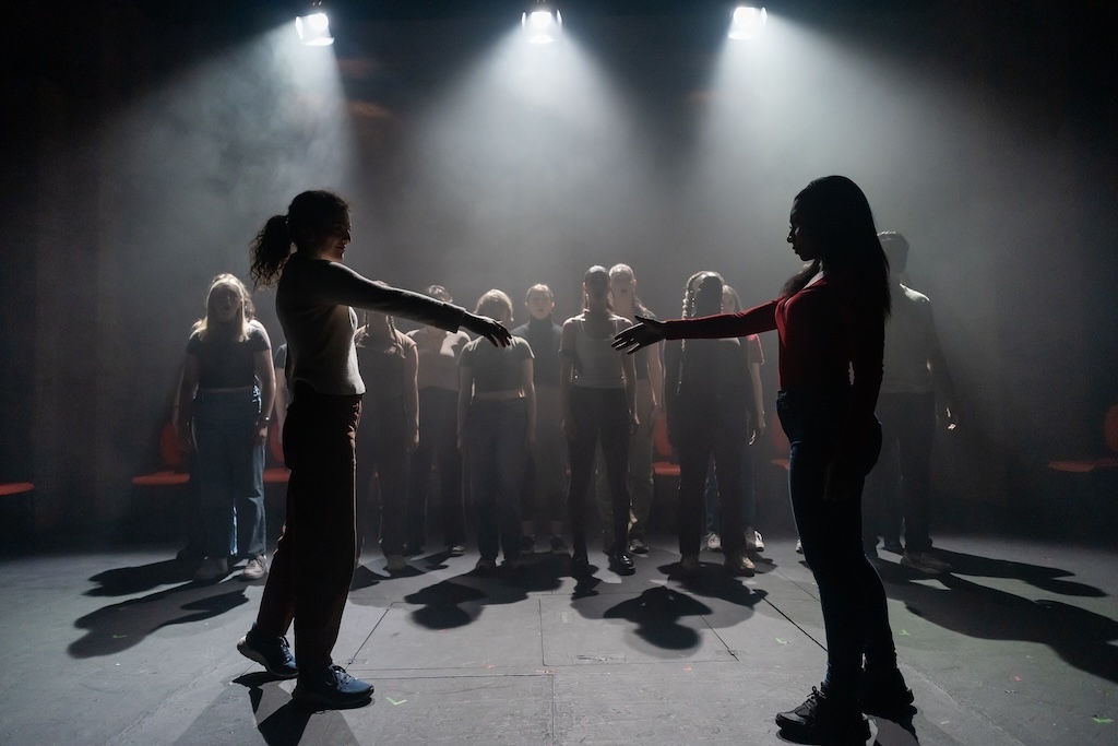 Action shot of the spring 2024 Shakespeare in Performance at RADA final presentation of Julius Caesar. Two actors, backlit, face each other on stage with arms outstretched. Behind them, a group of actors stands in mid-performance.